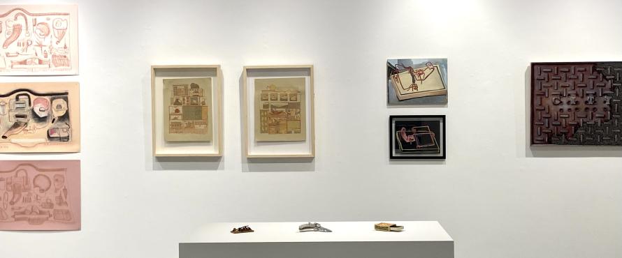 Gallery view of multiple artworks displayed on a wall, prints, drawings, paintings, and a pedestal with small ceramic objects that look like a mouse trap, matchbox and pocket knife