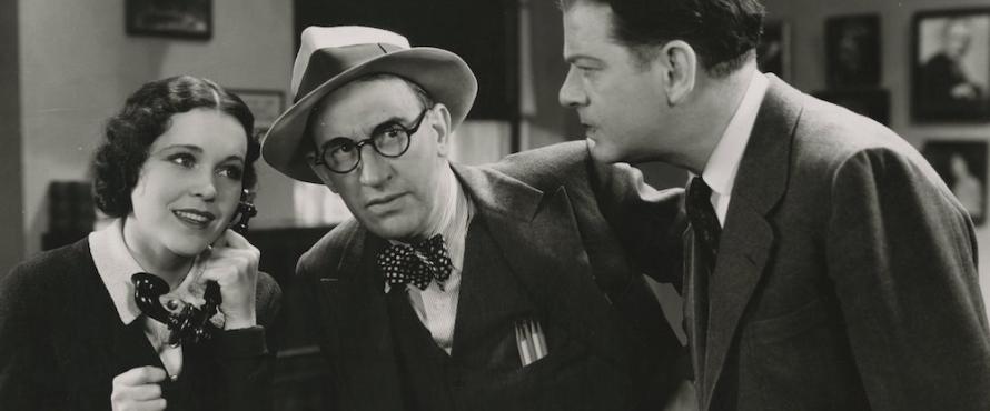 Still frame from the film "Okay America!" showing two men listening to a woman who is using the telephone.