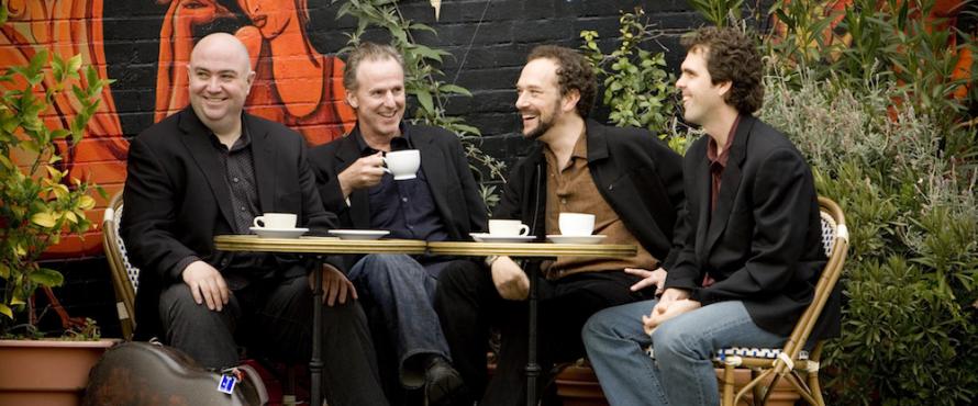 The four male members of the Los Angeles Guitar Quartet sitting at a table, drinking coffee and smiling with a colorful mural and plants in the background