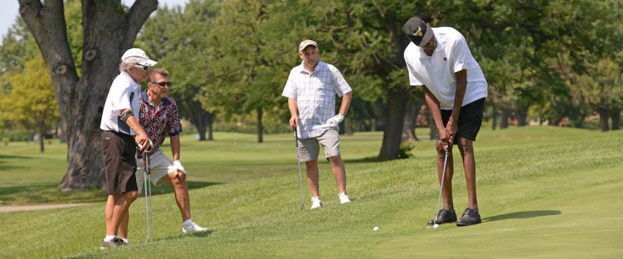 Three men watch as another man putts a golf ball on a golf course