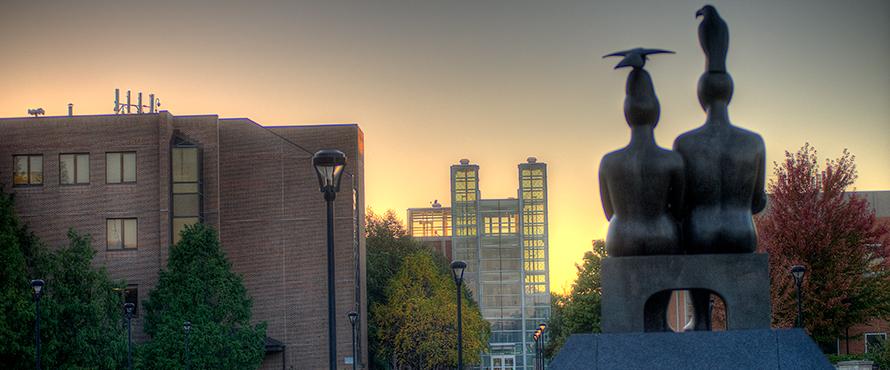 The "Serenity" statue on the University Commons as seen from behind with the Library and Parking Facility in the background against a sunset sky