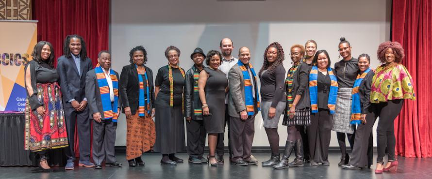 Sixteen people pose as a group on a stage at Carruthers Center for Inner City Studies