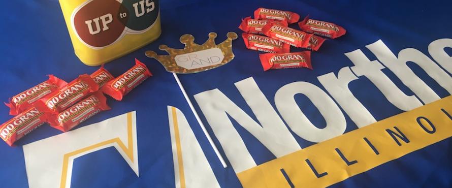 An Up to US branded yellow plastic box rests on an NEIU branded tablecloth amid a scattering of 100 Grand chocolate bars