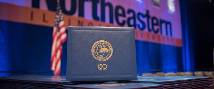 A diploma cover on display at December 2017 Commencement with an American flag and a branded Northeastern banner in the background