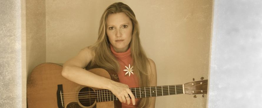 Rebecca Frazier, wearing a sleeveless orange shirt with a white flower on it, sits holding a guitar