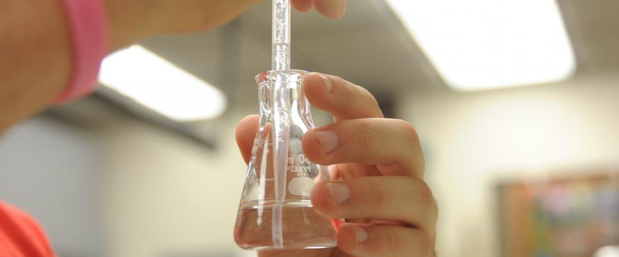 A pair of hands holding a glass beaker with clear fluid in it and a glass tube