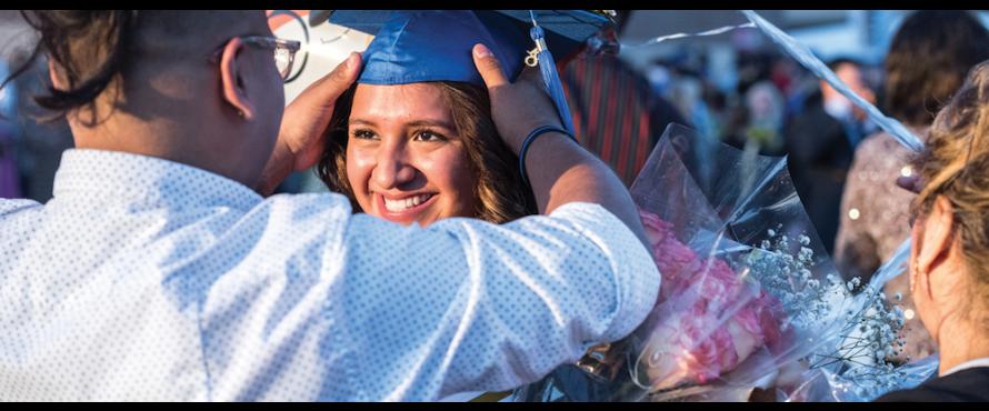 A person adjusts the blue mortarboard on the head of a smiling female graduate