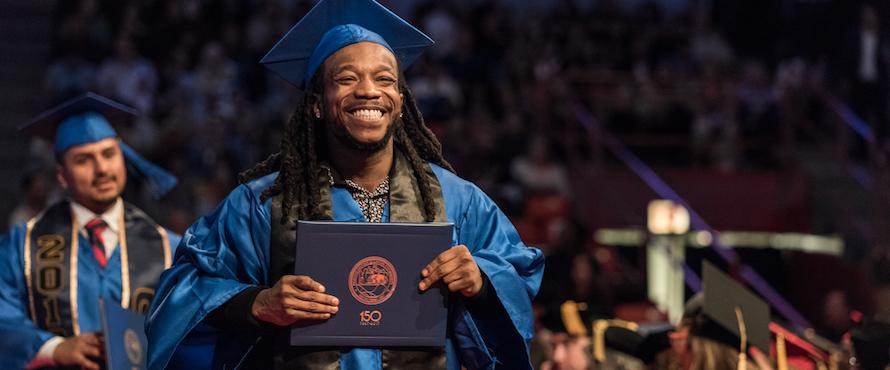A smiling male graduate wearing blue regalia holds his diploma 