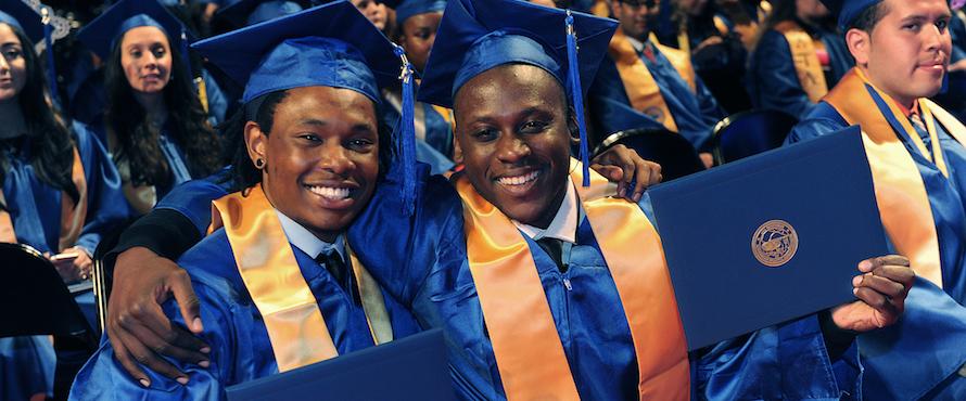 Two male graduates wearing blue caps and gowns and yellow stoles smile at the camera