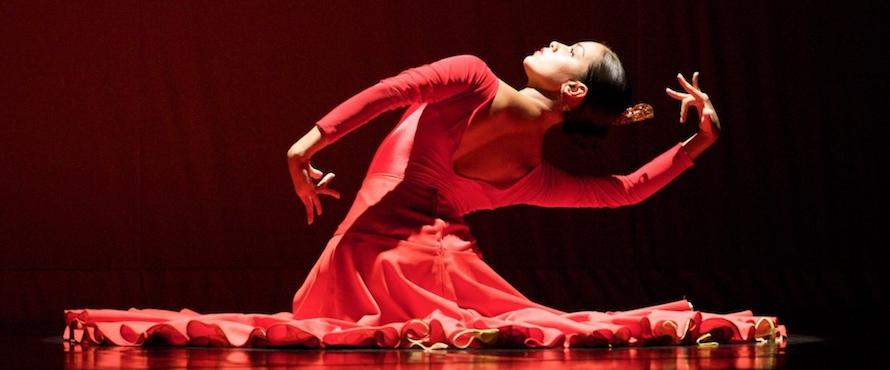 A female dancer wearing a red dress poses against a black background