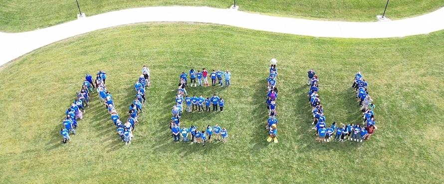 Students in blue T-shirts spell out NEIU in the grass.