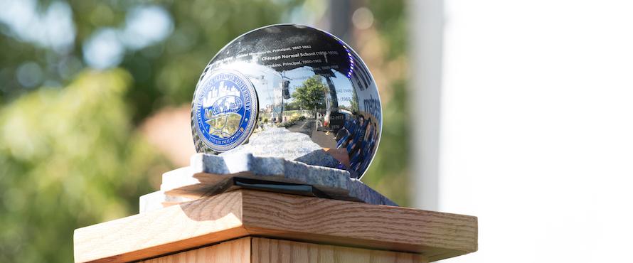 The metal, inscribed University Orb rests atop a wooden pedestal