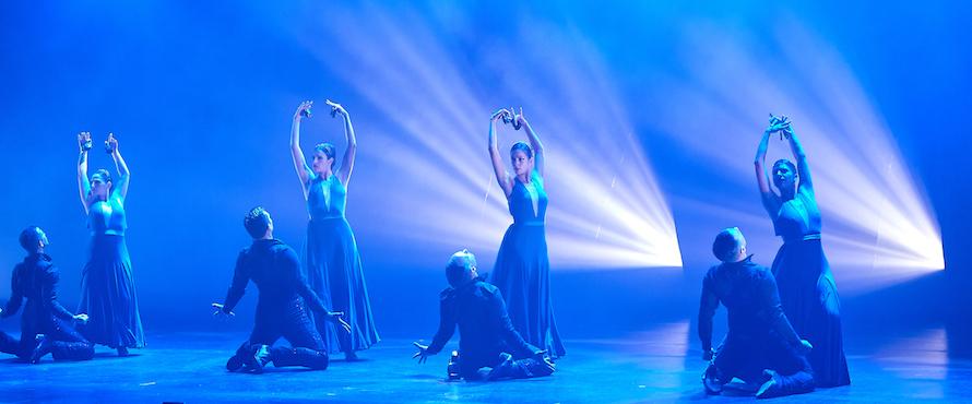Eight dancers strike poses on stage under blue lighting