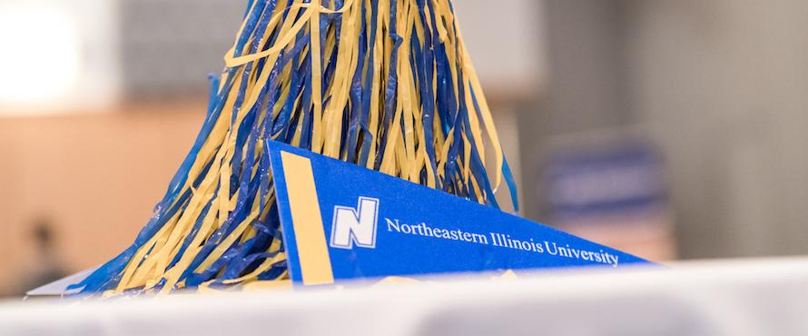 An NEIU pennant lies in front of a blue and gold pompom