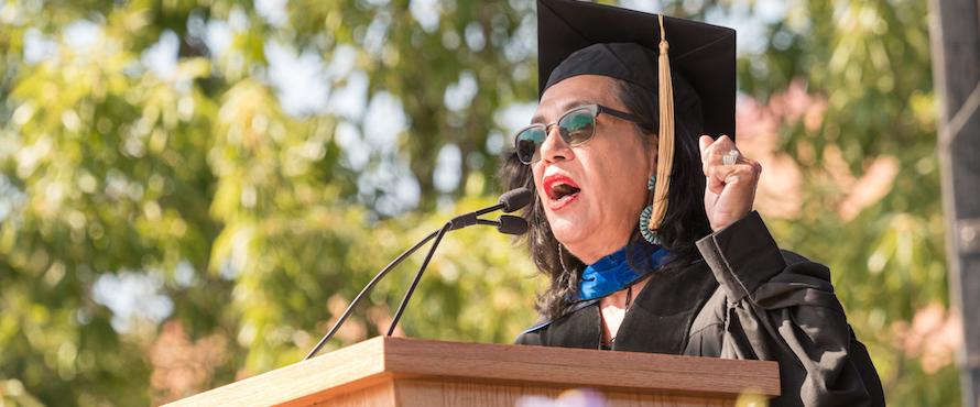 Ana Castillo wears academic regalia and stands outdoors at a dais as she addresses an audience