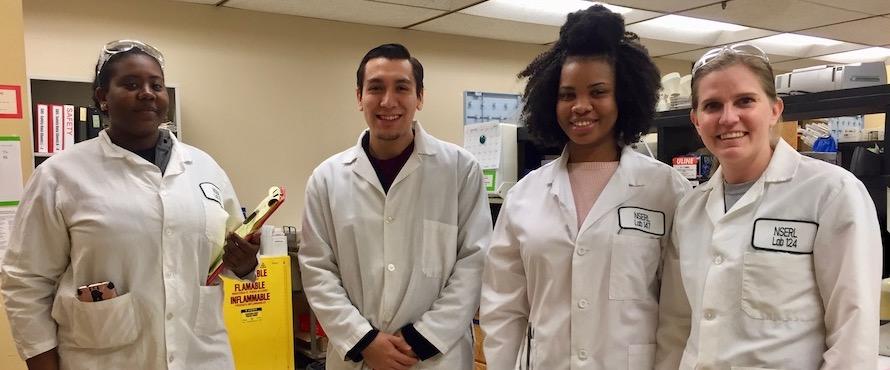 Four smiling students wearing white lab coats stand side by side in a laboratory