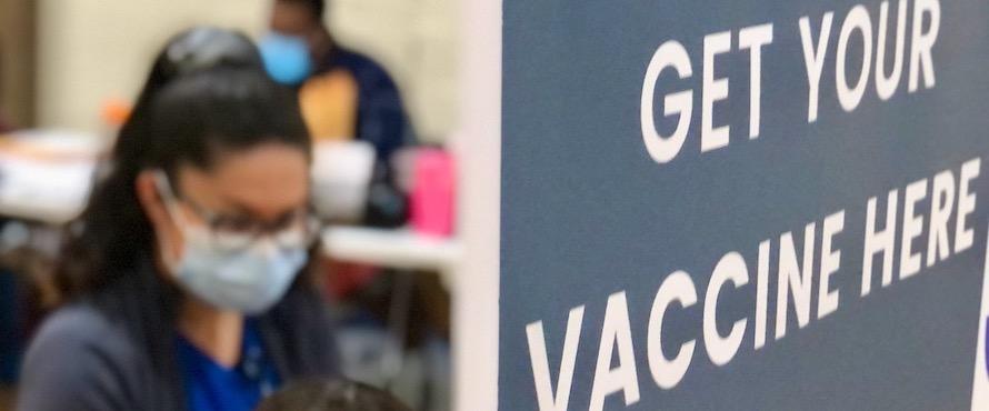 Photo of a person wearing a mask near a sign that reads, "Get your vaccine here" in Northeastern's P.E. Complex