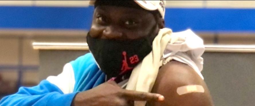 A man wearing a mask and baseball hat points to a bandage on his arm after getting a shot
