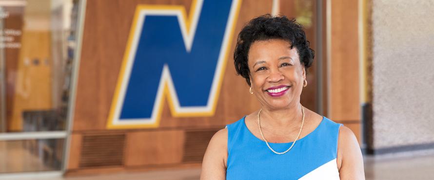 President Gibson wearing a blue top stands in front of a wall featuring NEIU's Flying N icon