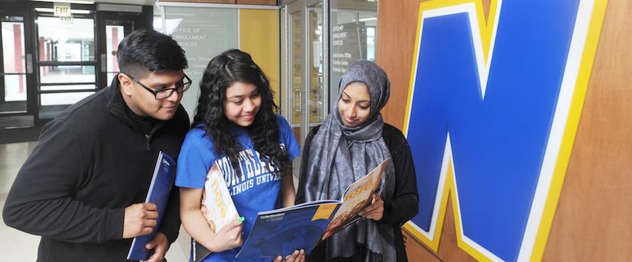 Three students look at a brochure while standing in front of a large blue N