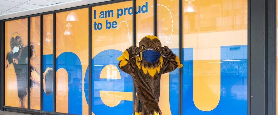 A photo of NEIU mascot Goldie wearing a mask in front of an I am proud to be NEIU sign