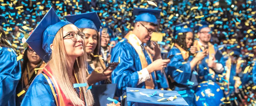 Graduates wearing blue regalia smile as confetti showers down around them