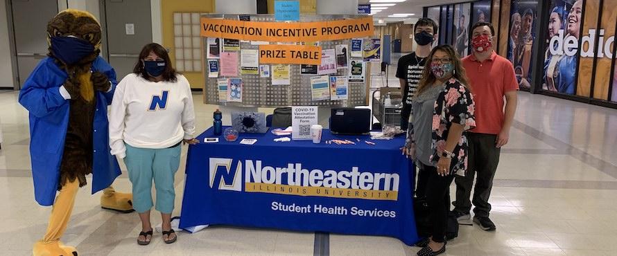 Photo of NEIU students and staff with Goldie at the Vaccine Incentive prize table