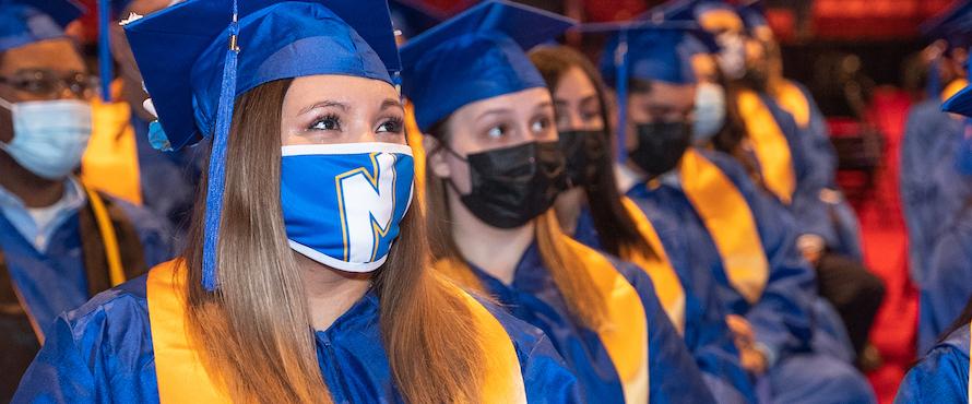 Photo of 2021 Northeastern graduates at Commencement wearing blue caps and gowns and yellow stoles. The student in the foreground is wearing a Northeastern face mask, while others wear black face masks. 
