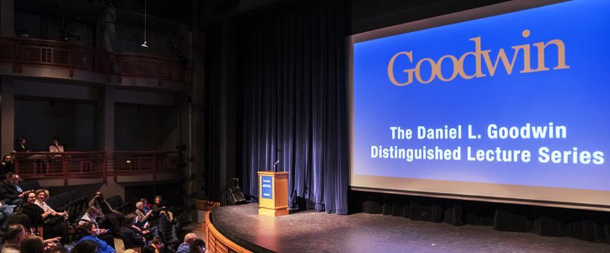 Photo of the NEIU Auditorium on the Main Campus with a large screen with the text "The Daniel L. Goodwin Distinguished Lecture Series" in white on a blue background. 