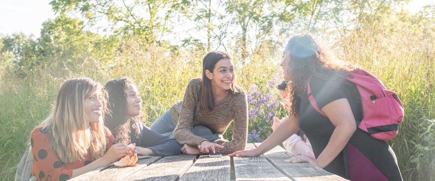 Five women gathered around an outdoor picnic table smile and laugh together
