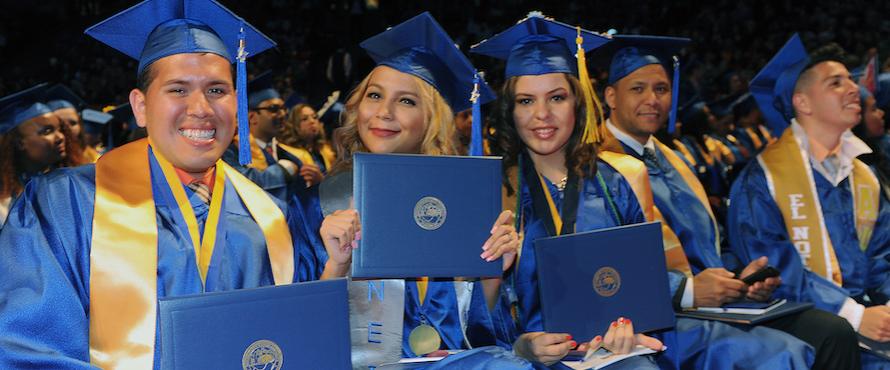 Smiling graduates in blue caps and gowns sit side by side at Commencement