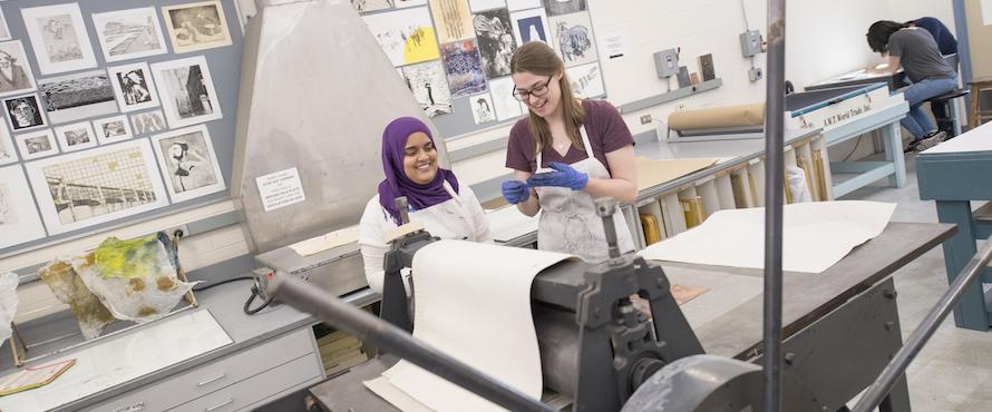 Two students wearing white smocks smile as they stand in the printmaking studio