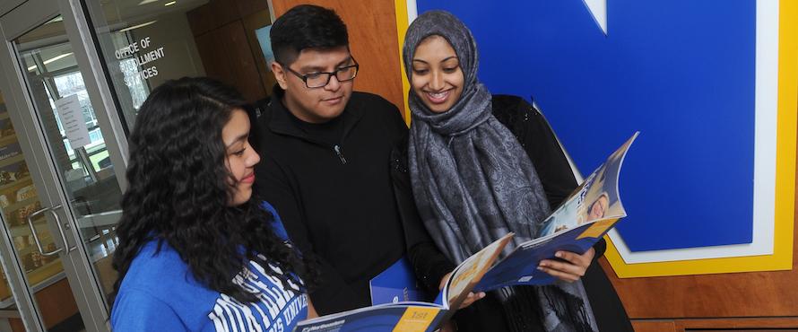 Three students look at an NEIU enrollment booklet outside of the NEIU enrollment office in front of the flying N.