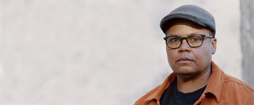 Photo of Keeanga-Yamahtta Taylor wearing a black t-shirt with orange button-down top, gray hat and black-rimmed glasses
