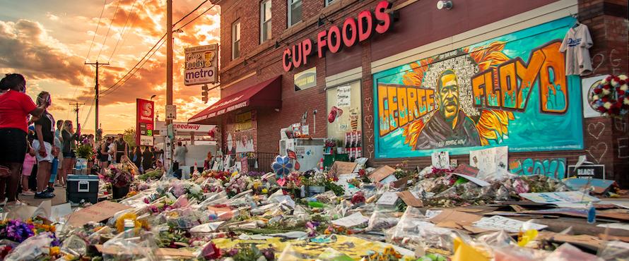 Photo of the mural tribute to George Floyd with flowers cards and other signs laid in front outside of Cup Foods in Minneapolis, Minnesota as mourners pay respects in June 2020. Photo by Vasanth Rajkumar/Wikimedia Commons. 