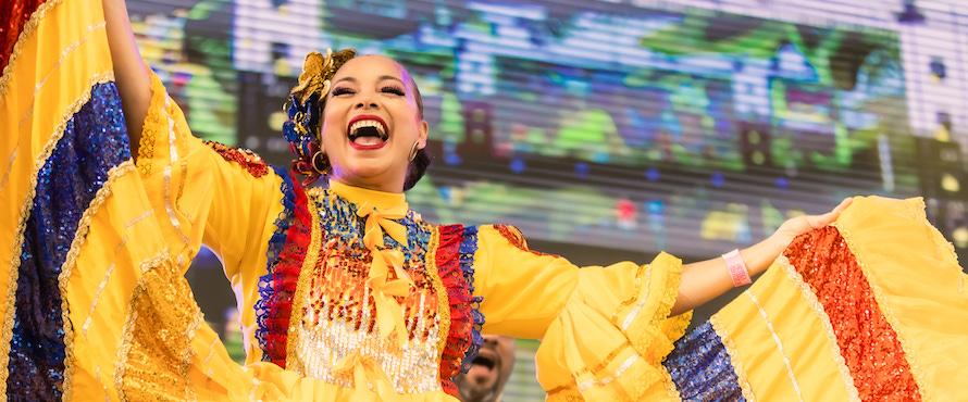 A photo of a dancer in a bright yellow, blue and red sequin folkloric dress with her holding dress ends outstretched in a dance pose, smiling, during Colombia Fest Miami 2021. Photo by Colombian Fest Worldwide.