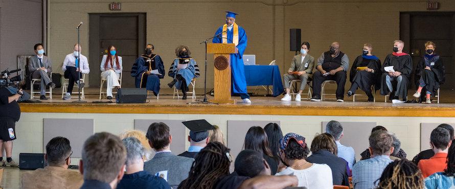 Northeastern Illinois University alumnus Michael Bell (B.A. '22 University Without Walls), speaks at a podium during his graduation ceremony at Stateville Correctional Center in October 2022. He is surrounded by professors and guest speakers sitting on a stage with audience members facing him in the foreground. Photo courtesy of Prison + Neighborhood Arts/Education Project.