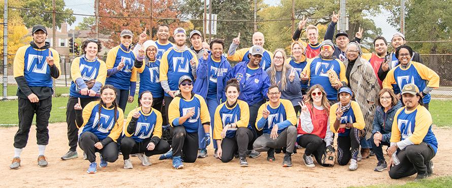 28 faculty and staff who played in the inaugural Joy and Wellness Softball Tournament wear their blue and yellow softball jerseys and smile for a group photograph at the NEIU baseball field.