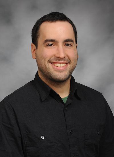 Adam Messinger wearing a black button up shirt sits in front of a gray backdrop