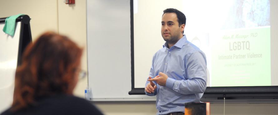 Adam Messinger wearing a light blue buttoned stands in front of a pull-down screen in a classroom setting