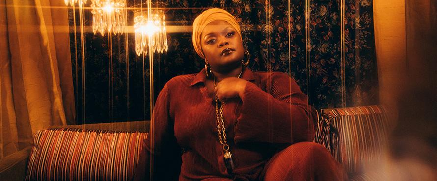 Kyshona wearing a maroon outfit, sitting on a striped couch, against a floral background. The photo is accented in warm lighting tones. A few chandeliers are also visible in the background. Photo by: Anna Haas