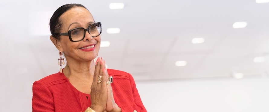 A photo of Murrell J. Higgins Duster wearing black glasses and a red dress with red blazer. Her hands are pressed together in a symbol of gratitude.