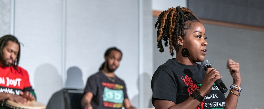 A person speaking at a past Northeastern Black History Month event, holding a microphone. Two other people are visible in the background playing drums. The speaker is wearing a black shirt with red, white and green graphics. The text on the shirt reads "Celebrating Black Resistance: Black History Month." 