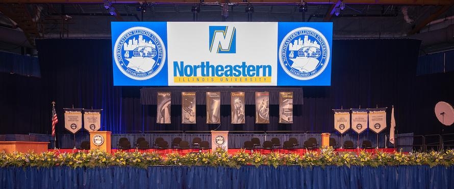 A photo of the stage at a Northeastern Illinois University Commencement Ceremony, set with the flags of the United States, State of Illinois, the University colleges and University Values. The Presidential Seal is visible on a large video screen and two podiums.