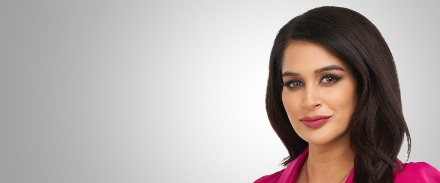 A headshot photo of PJ Randhawa wearing a pink blazer, standing against a gray background.