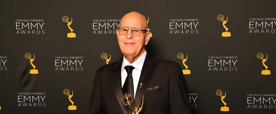 A photo of Steve Novak wearing glasses, a black suit, white collared shirt and black tie, smiling, holding an Emmy Award against a backdrop for the Chicago/Midwest Emmy Awards. 