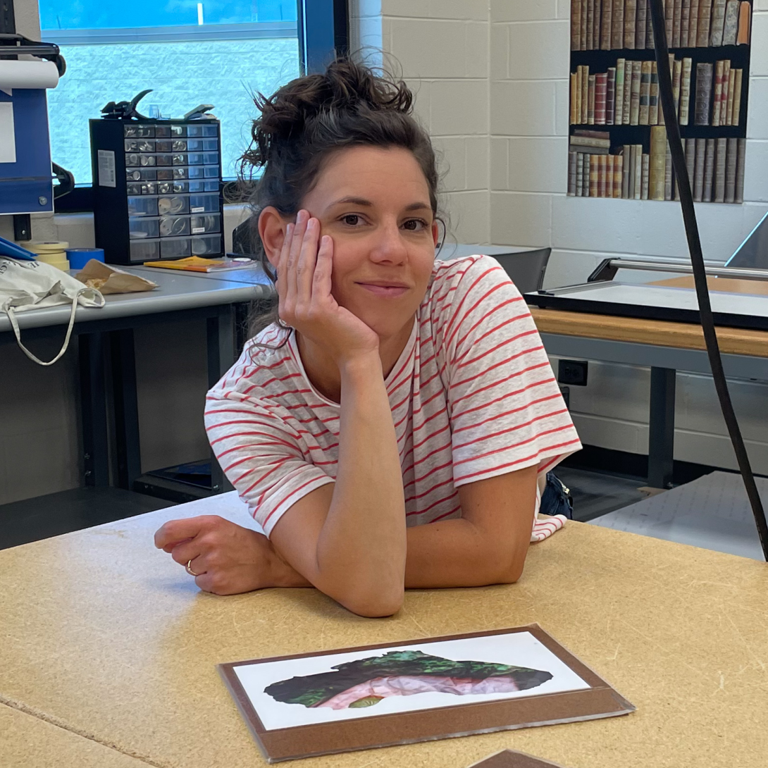 Candice looking at camera with one arm resting on face, leaning on table in front of an artwork printed on white paper placed on a cardboard rectangular backing