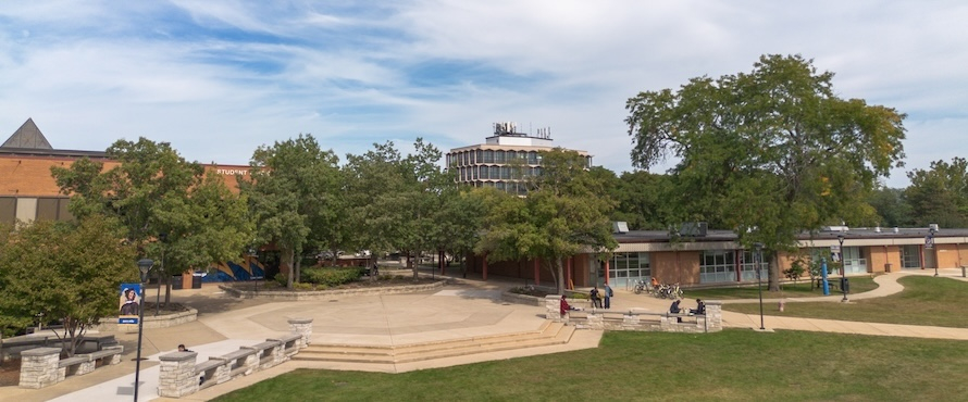 A photo of Northeastern Illinois University's Main Campus on a sunny day. The Student Union, Building B and Jerome M. Sachs Administrative Building are visible among trees and people sitting on the University Commons.    