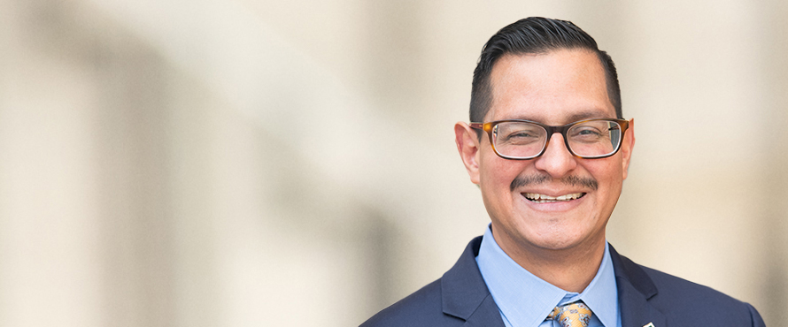 A headshot photo of Terry C. Mena, Ph.D., wearing a navy blue suit, blue collared shirt and yellow tie and glasses, smiling. 