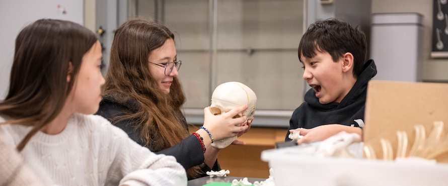 A photo of three young students participating in the 2025 Chicago River Student Congress. One is holding a model of a human skull. Another is looking at the skull as if in amazement.